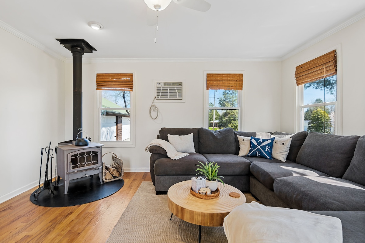 A comfortable living area is highlighted by a dark sectional sofa, positioned near a modern wood stove. Natural light is allowed in through windows with bamboo shades, and a round coffee table with decorative items sits on a neutral-colored rug.
