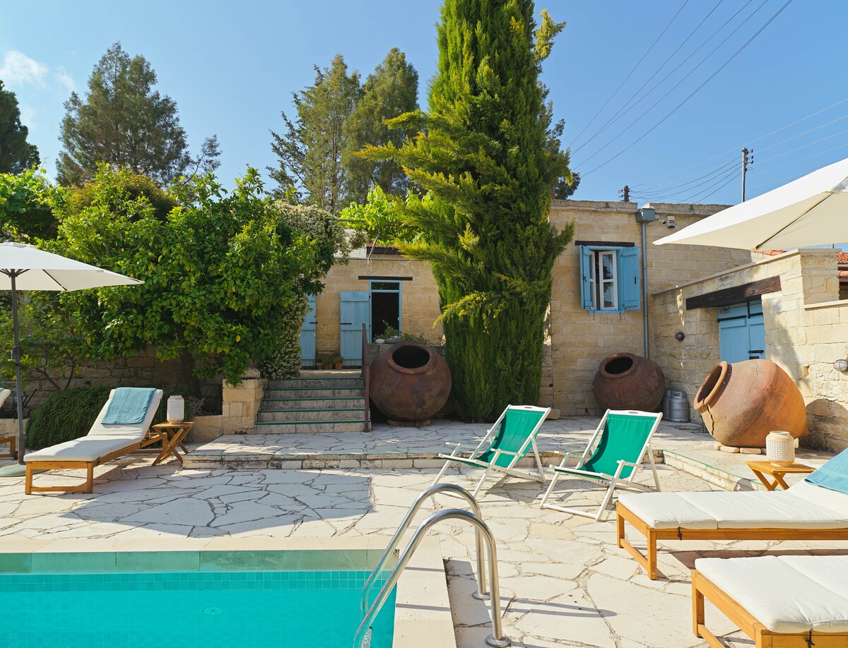 The outdoor area features a serene swimming pool surrounded by sun loungers and umbrellas. Lush greenery lines the space, with stone steps leading to a charming stone structure and two large clay pots. Light filters through the trees, enhancing the tranquil setting.