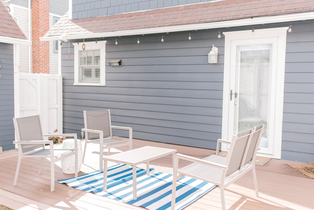 An outdoor patio area features light-colored furniture arranged around a rectangular table. A striped rug adds a touch of color to the space. String lights are hung above, creating a cozy ambiance, while a door leads into the interior of the cottage.