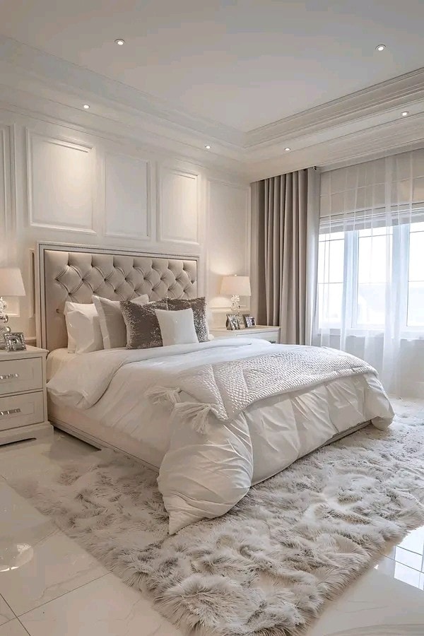 A stylish bedroom features elegant paneling and a plush, oversized bed adorned with decorative pillows. Light neutral tones are complemented by soft textures, including a fluffy area rug. Gentle lighting enhances the serene atmosphere near a large window draped with sheer curtains.
