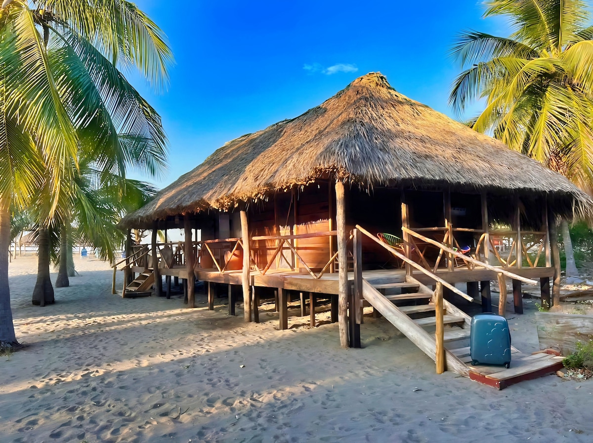 A thatched-roof structure is elevated on wooden stilts, situated on sandy terrain. Balconies with railings provide shaded areas beneath the roof. Lush palm trees frame the scene, while a clear blue sky enhances the serene coastal atmosphere.
