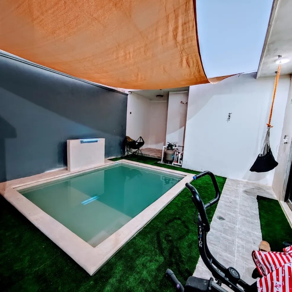 A small, serene pool is surrounded by textured tile flooring and artificial grass. The area is shaded by a canopy, providing a comfortable retreat. A pair of folding chairs and a hammock are positioned nearby, inviting relaxation in the minimalist space.
