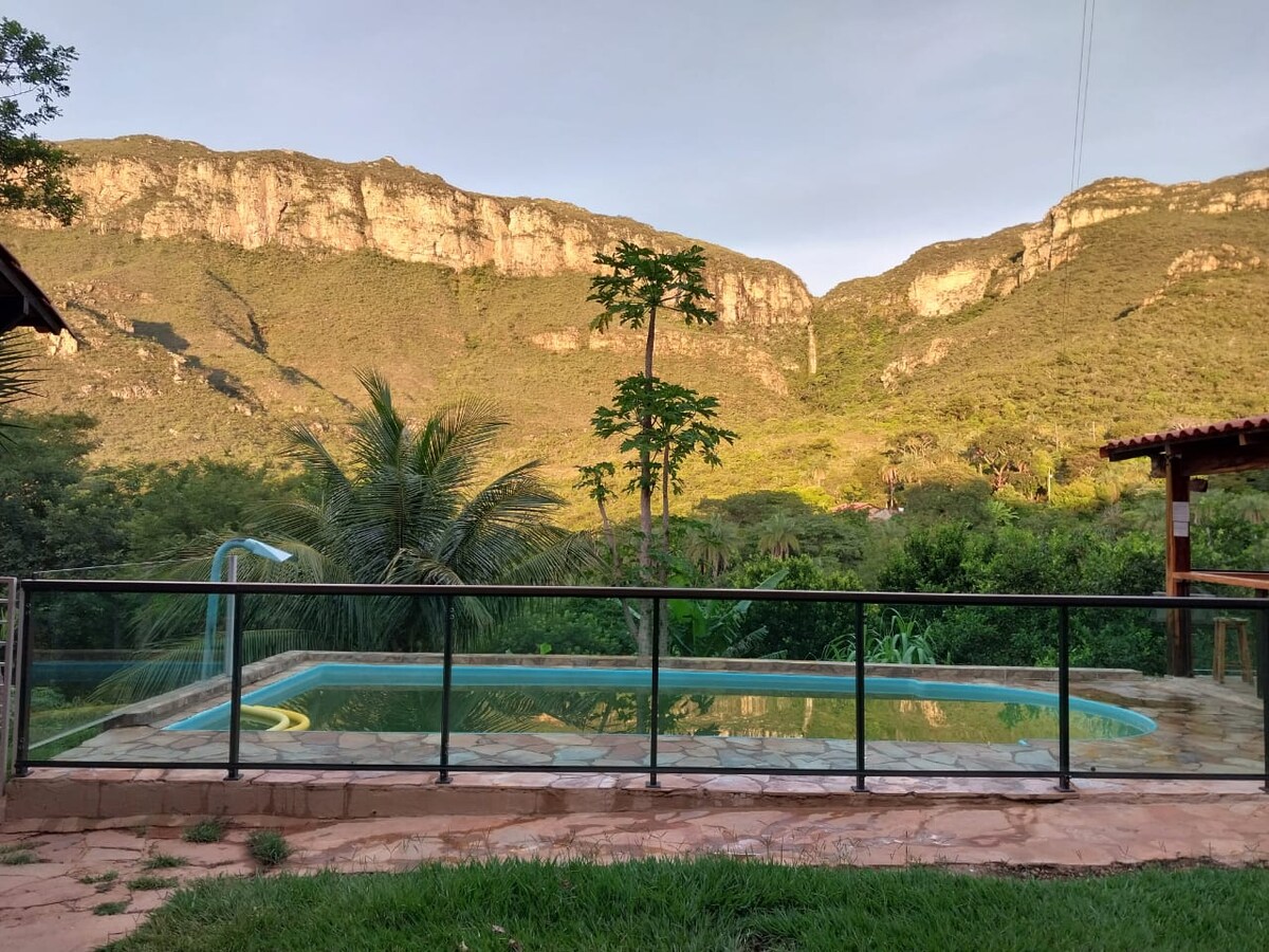 A refreshing pool is surrounded by a stone patio, with lush greenery in the foreground. In the background, majestic mountains rise, showcasing a range of natural hues under the soft light. A sense of tranquility is evident in this outdoor space.
