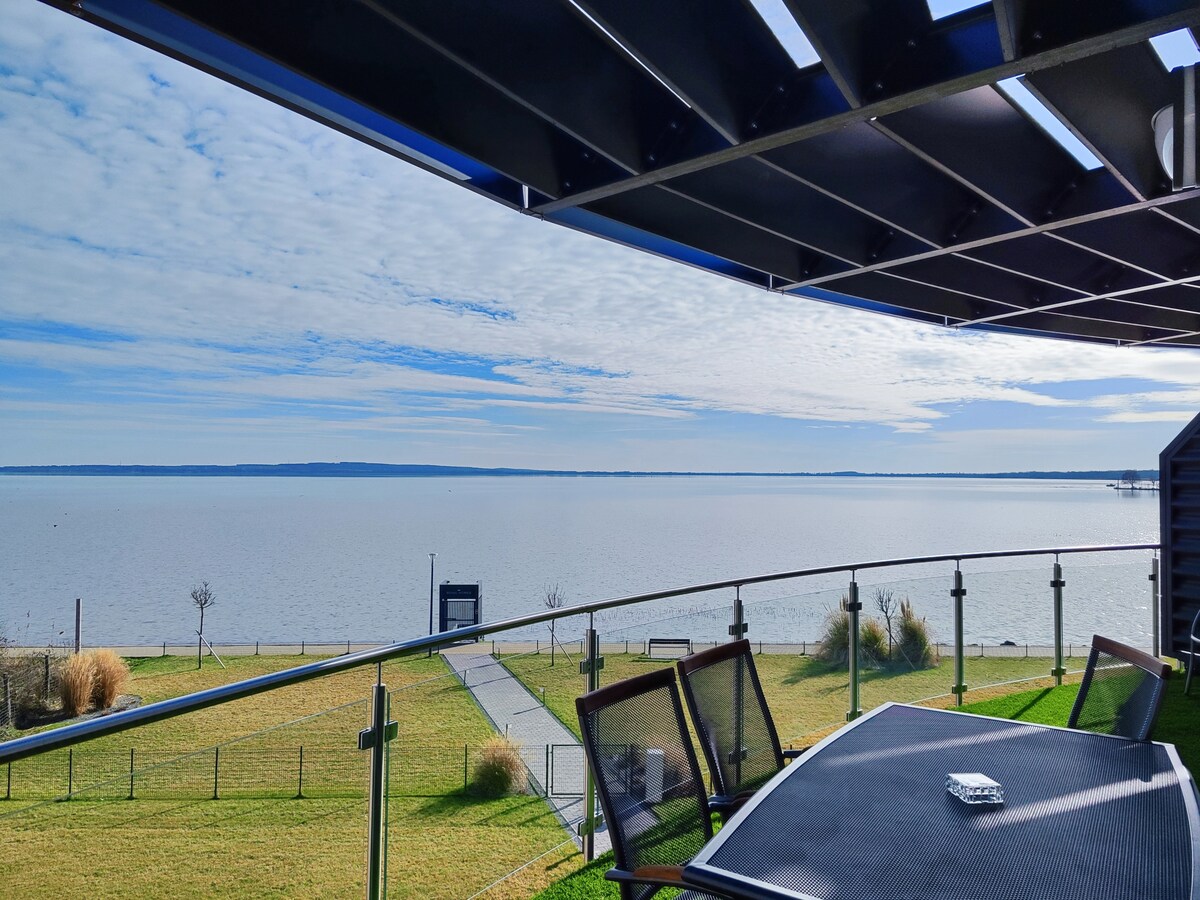 A spacious balcony offers a panoramic view of the serene water, framed by soft clouds and grassy shores. Outdoor furniture includes a table and several chairs positioned to enjoy the landscape, which features a pathway leading towards the shoreline.