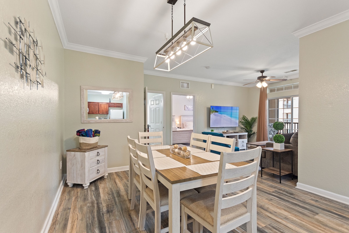 A dining area features a wooden table with six chairs, set with neutral table runners. A mirror is mounted on the wall, reflecting light, and a decorative piece is displayed nearby. The space opens to a living area, showcasing a TV and access to the balcony.