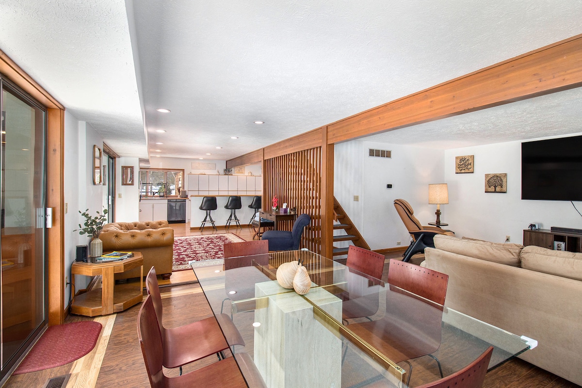 An open living area features a dining table set for six, surrounded by stylish chairs. A sectional couch and exercise equipment are visible in the background, while a large window allows ample natural light to brighten the space.