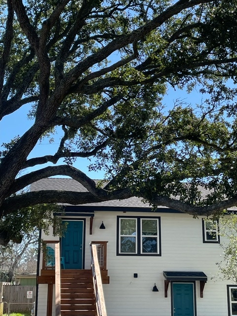 The exterior of the cottage is framed by a large oak tree, with a wooden staircase leading to a teal door. Bright blue skies are visible above, complementing the white siding and sleek black windows of the home.