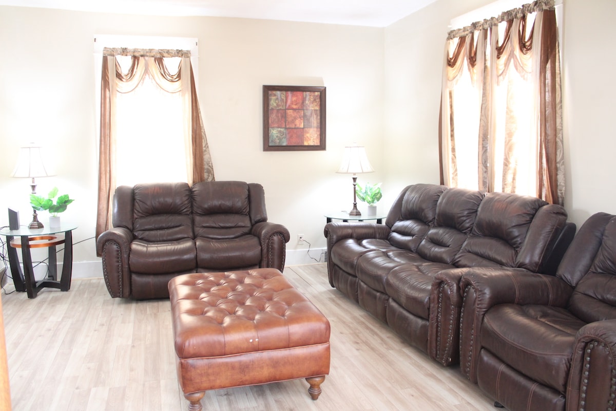 A living room is featured with two dark leather sofas and a button-tufted brown ottoman at the center. Natural light filters in through two windows adorned with flowing drapes. Two side tables with lamps sit beside the sofas, and decorative plants add a touch of greenery.
