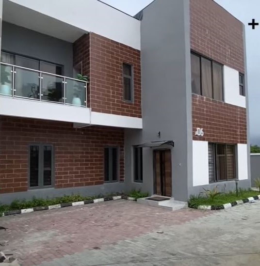 A modern, two-story terrace duplex is displayed, featuring a clean exterior with a mix of brick and grey walls. Large windows provide natural light, while a small balcony is positioned above the entrance. Paved pathways and greenery surround the entrance area.