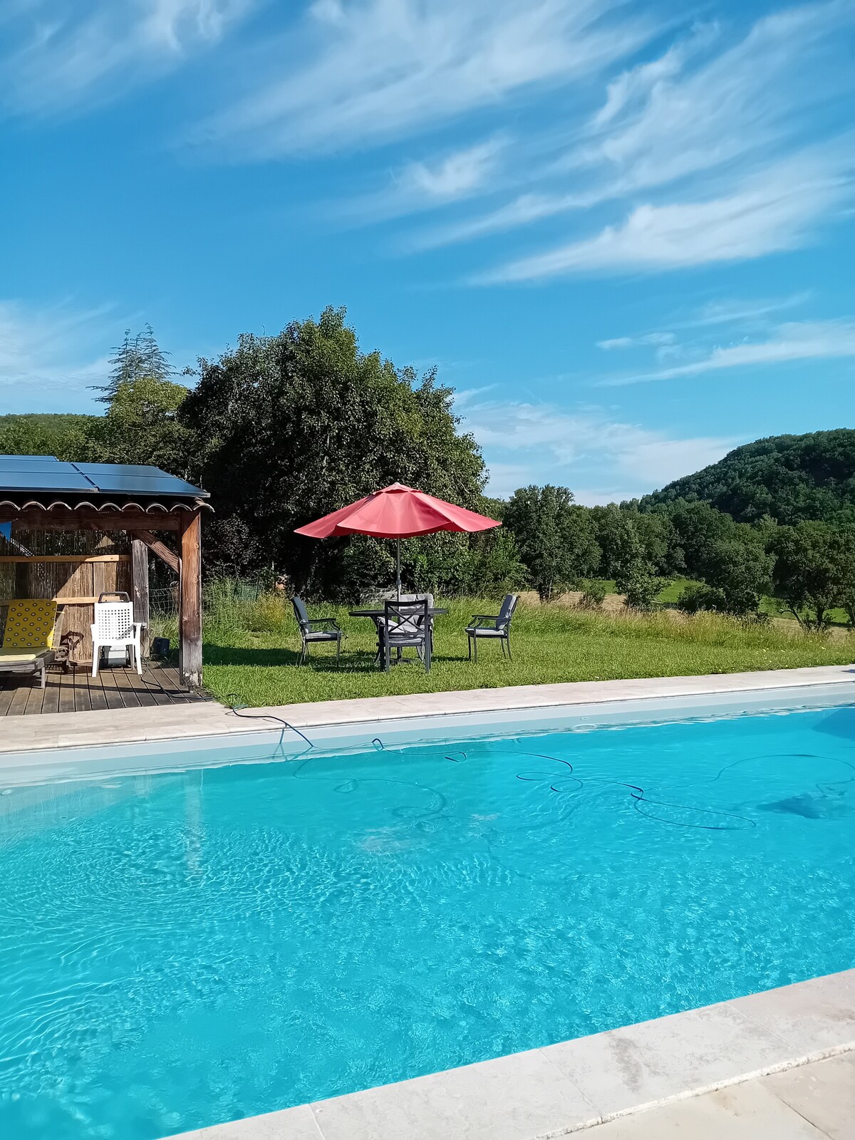 A swimming pool reflects the blue sky, accompanied by a shaded patio area featuring a red umbrella over a table and chairs. Lush greenery and rolling hills form the backdrop, enhancing the outdoor serenity of the space.