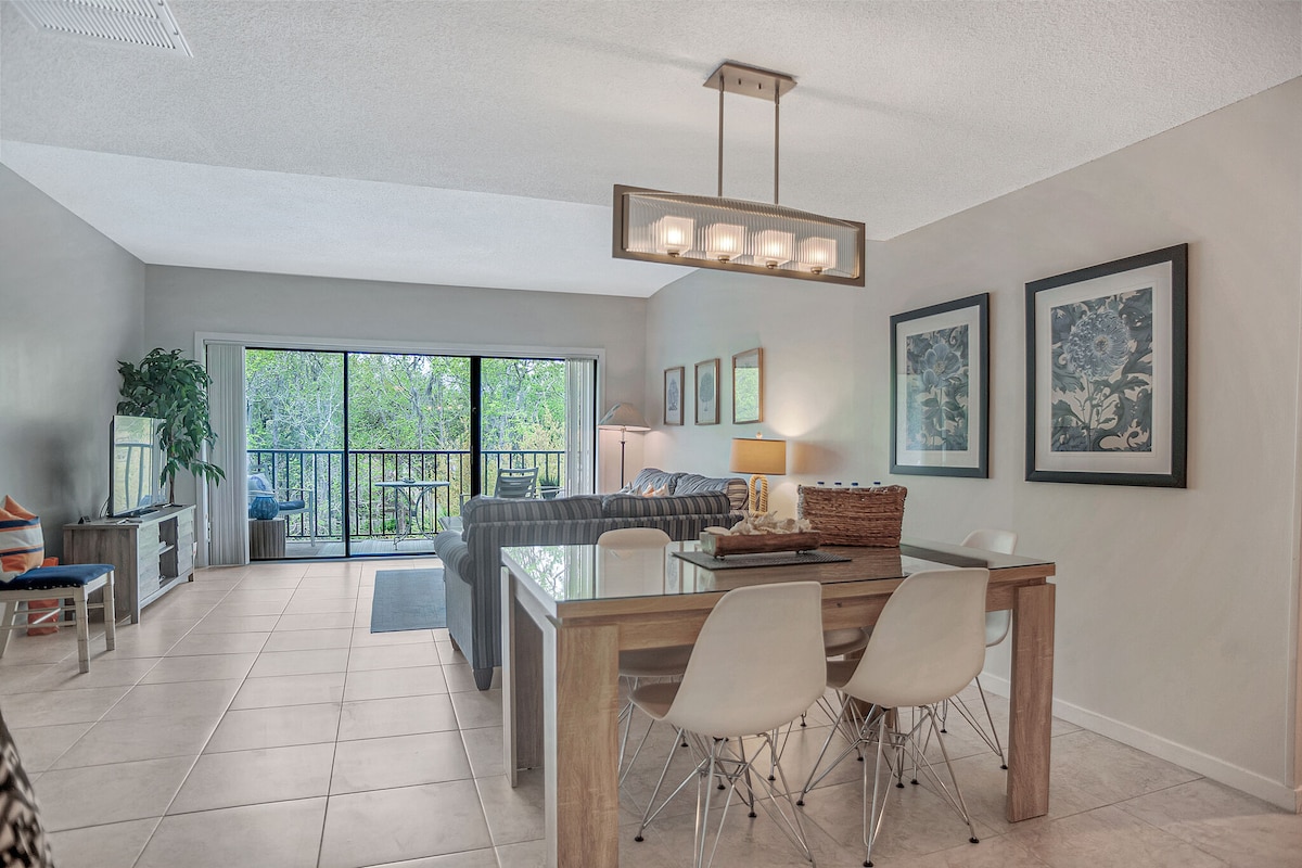 The open living and dining area features tile flooring and a rectangular dining table accompanied by white chairs. A modern light fixture hangs above the table, while a comfortable sofa sits in the background. Sliding glass doors lead to a balcony with views of greenery.