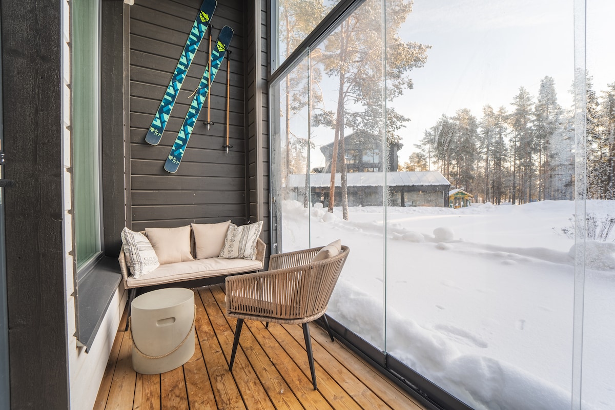 A glassed-in terrace features a round table and a wicker chair, providing a cozy seating area surrounded by snow-covered scenery. Ski equipment is displayed on the wall, emphasizing the winter atmosphere. The wooden floor adds warmth to the space.