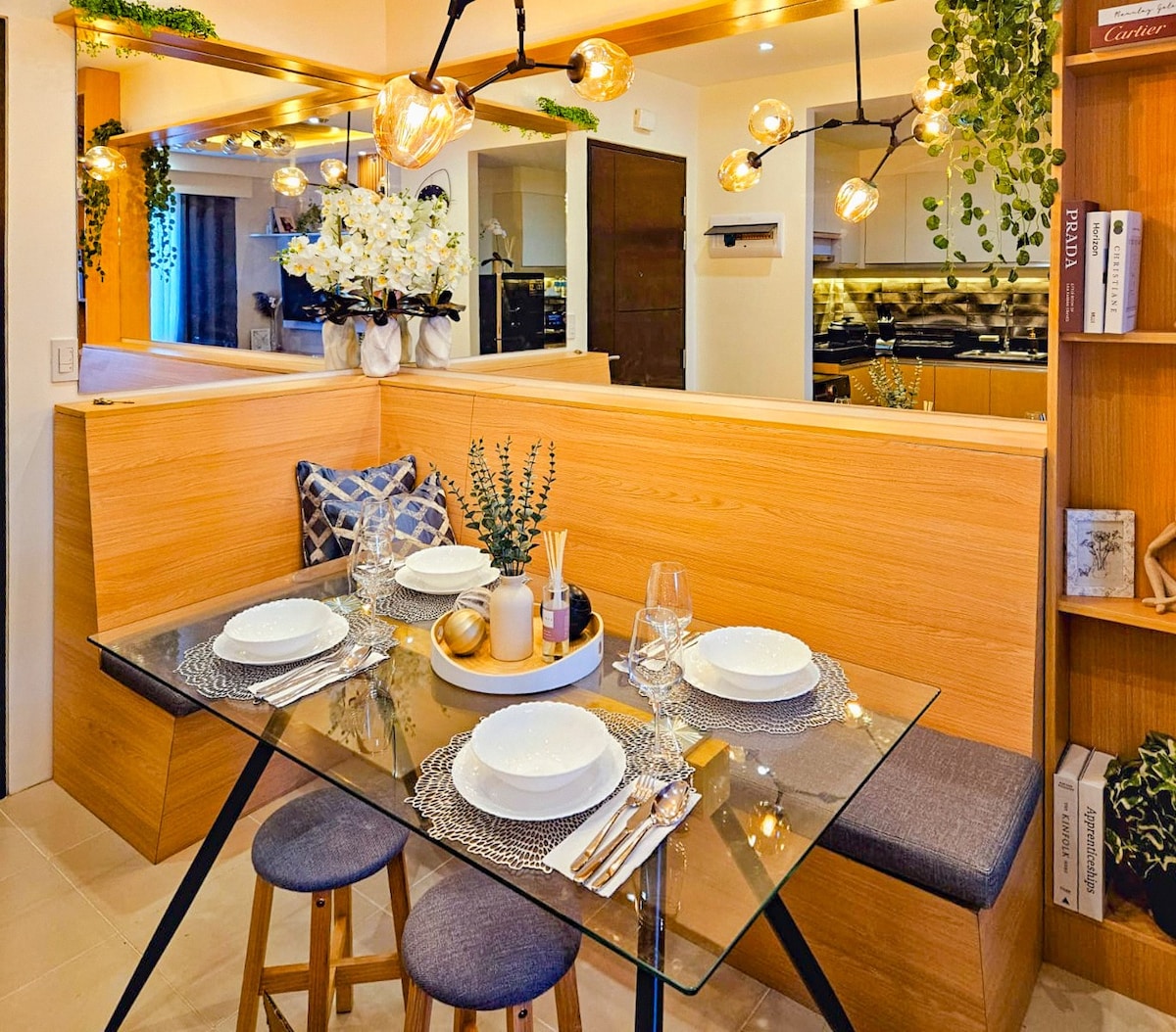 A glass dining table is set for four, featuring elegant tableware and decorative elements. Warm lighting highlights the wooden accents and green plants, creating a cozy dining area. A nook with cushions provides additional seating, contributing to an inviting space for shared meals.