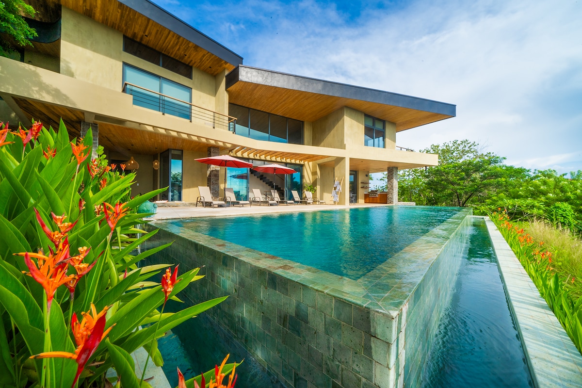 A modern villa is showcased from a landscaped perspective, featuring an inviting infinity pool that blends with the surrounding nature. Lush green foliage and vibrant flowers frame the scene, while expansive glass windows reflect the open sky and enhance natural light within the space.