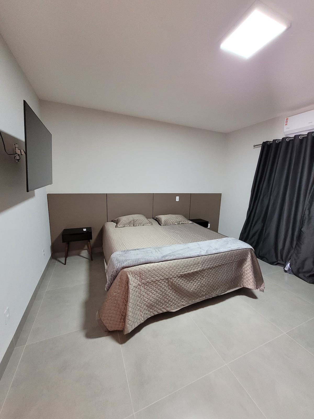 A spacious sleeping area is featured, showcasing a neatly made bed with neutral bedding. A small nightstand sits beside the bed, and a television is mounted on the wall opposite. Dark curtains frame a window, allowing soft light to enter the room.