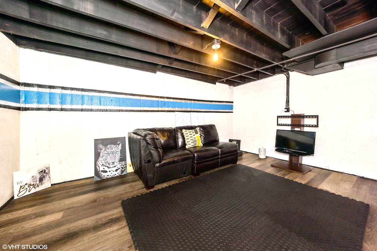 A cozy area is furnished with a dark leather couch, accented by a decorative pillow. A modern television is mounted on the wall, and artwork is displayed on the floor, complementing the simplicity of the space. The flooring is a dark, textured material beneath.