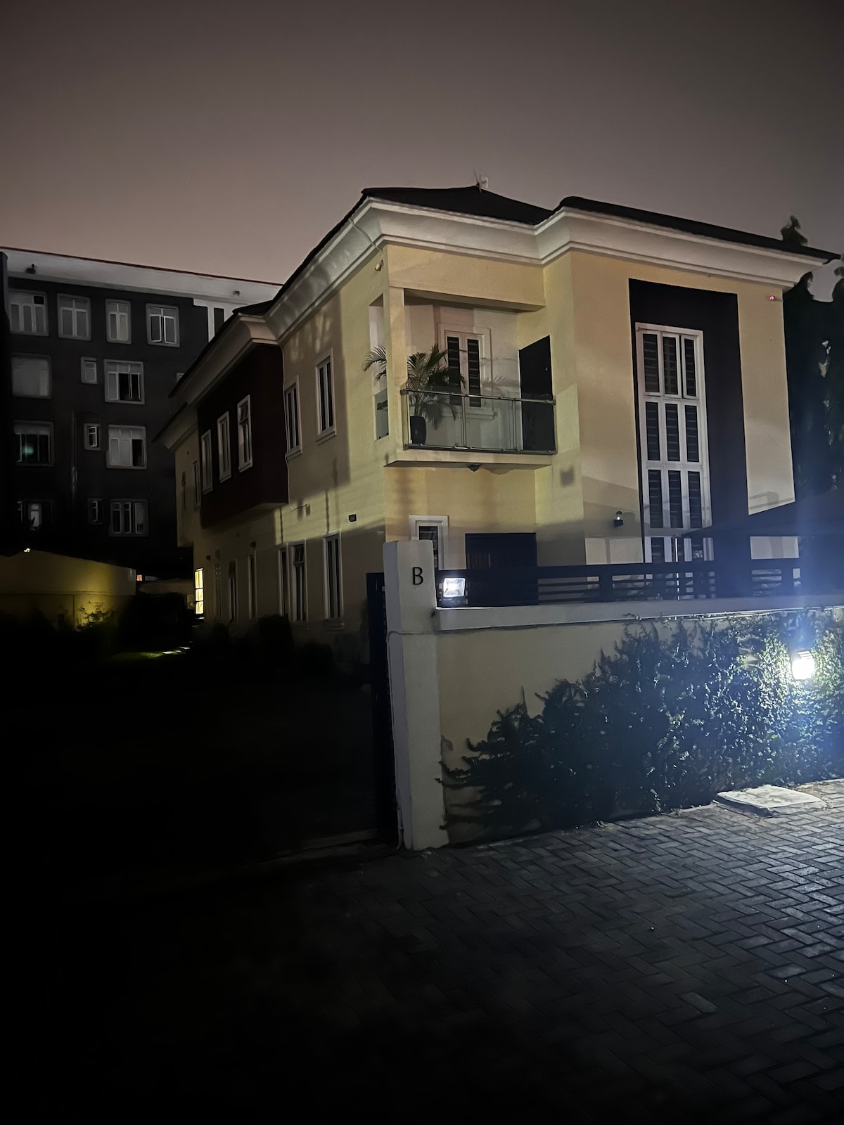 The exterior of a two-story residence is softly illuminated at night, highlighting its contemporary architectural features. The front façade displays a blend of light and dark colors, with a large entrance door flanked by vertical windows. The surrounding area includes hedges and paved walkways.