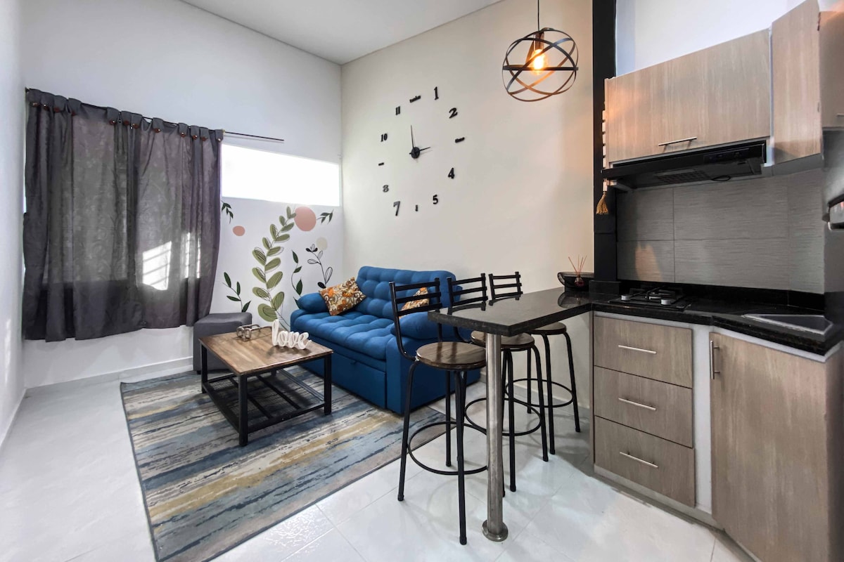 An inviting living area is presented with a blue sofa and a wooden coffee table resting on a patterned rug. A small dining bar with high stools complements the adjacent kitchen, which features modern cabinetry. Natural light filters through sheer curtains, enhancing the space.