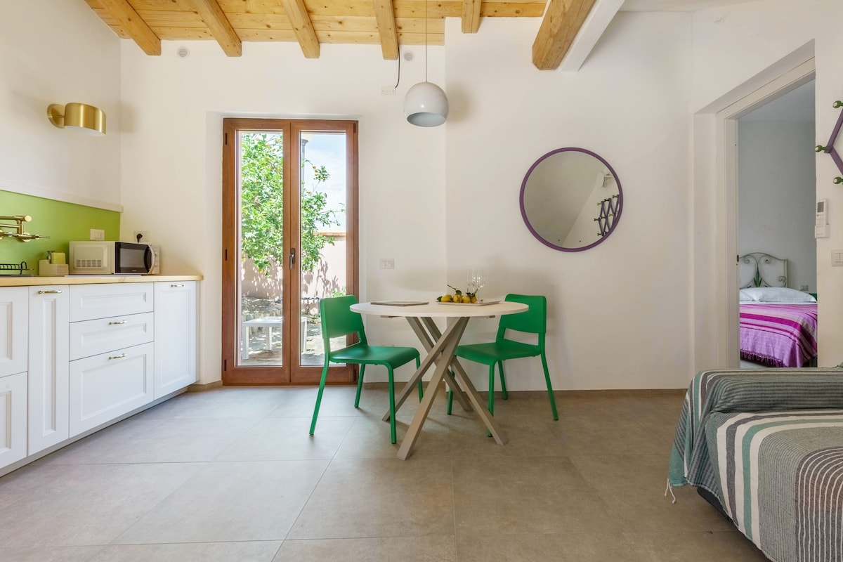 A bright dining area features a small round table with two vibrant green chairs. Natural light enters through the large glass doors, revealing a view of the garden. A functional kitchenette is designed in white, accompanied by a microwave and minimal decor.