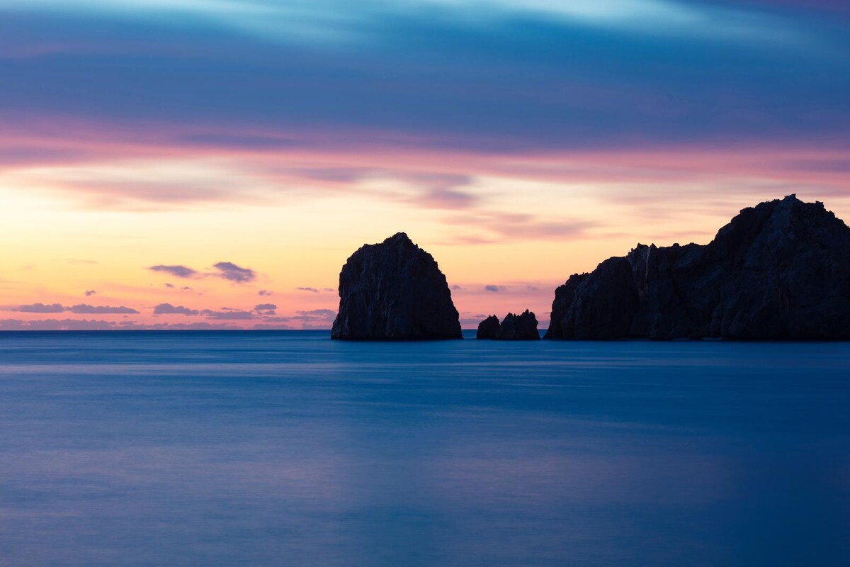 The tranquil waters of the Sea of Cortez reflect soft hues of orange, pink, and blue during sunset. Distinct rock formations are silhouetted against the colorful sky, creating a serene coastal scene. The gentle waves caress the shoreline, enhancing the peaceful atmosphere.