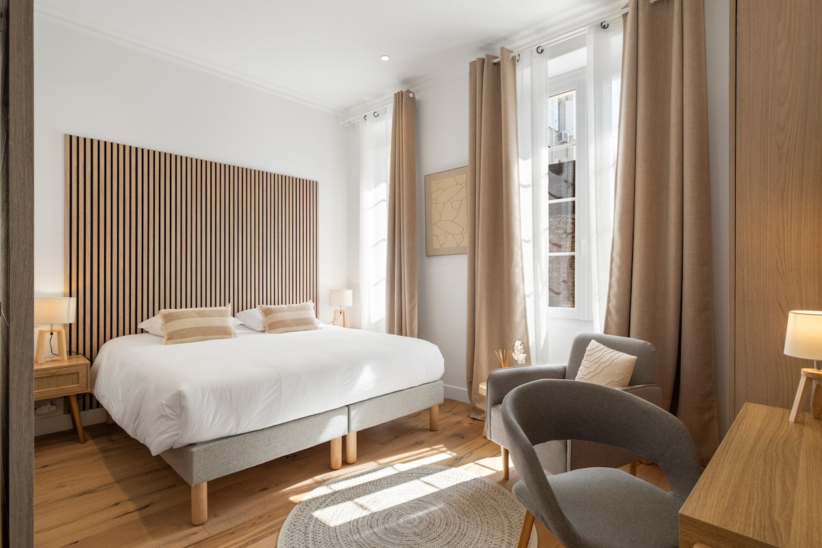 A bright and airy bedroom features a spacious bed with a wooden headboard. Natural light streams through large windows dressed with light curtains, highlighting the warm wood flooring. A comfortable chair, a round rug, and a simple desk add to the cozy ambiance.