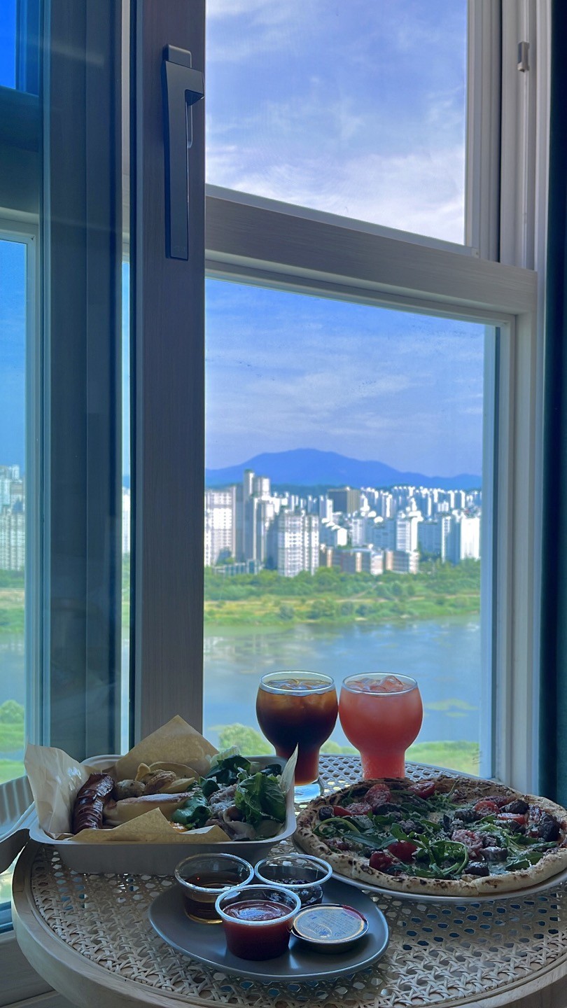 A dining table is set near a large window, showcasing a view of the city skyline and river below. Two colorful drinks and a plate of food, including salads and sauces, are arranged on a woven table surface, inviting a relaxed dining experience.