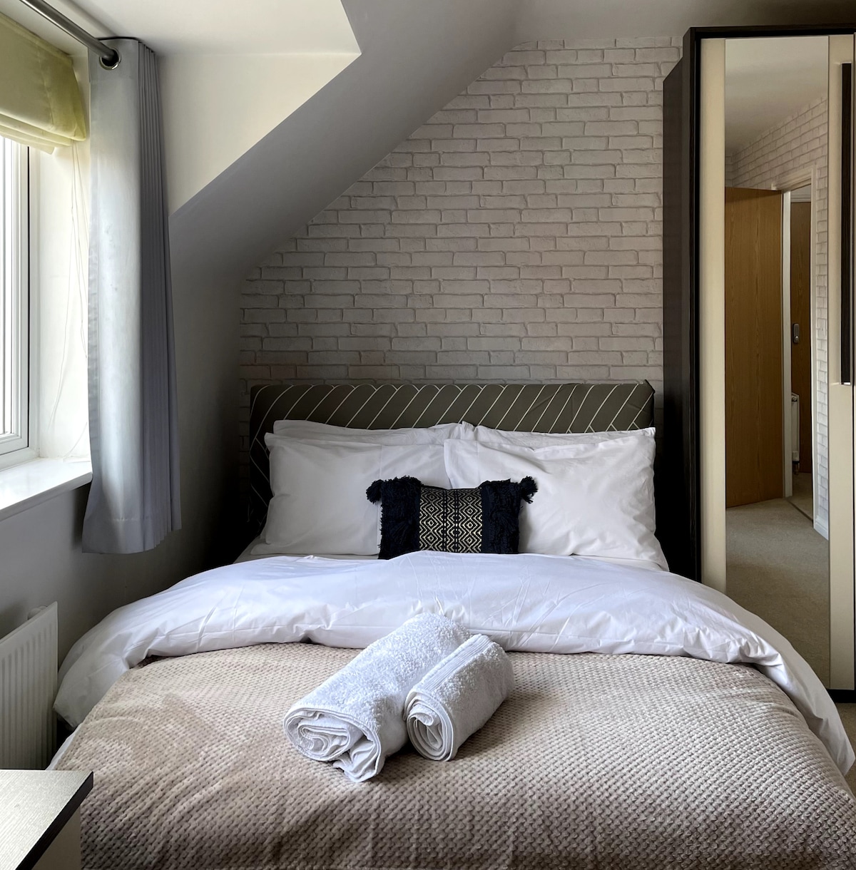 The master bedroom features a spacious bed with crisp white linens, complemented by a decorative pillow. Soft natural light enters through a window, while a neatly arranged pair of towels rests on the bed. A mirrored wardrobe is positioned nearby, reflecting the serene ambiance.