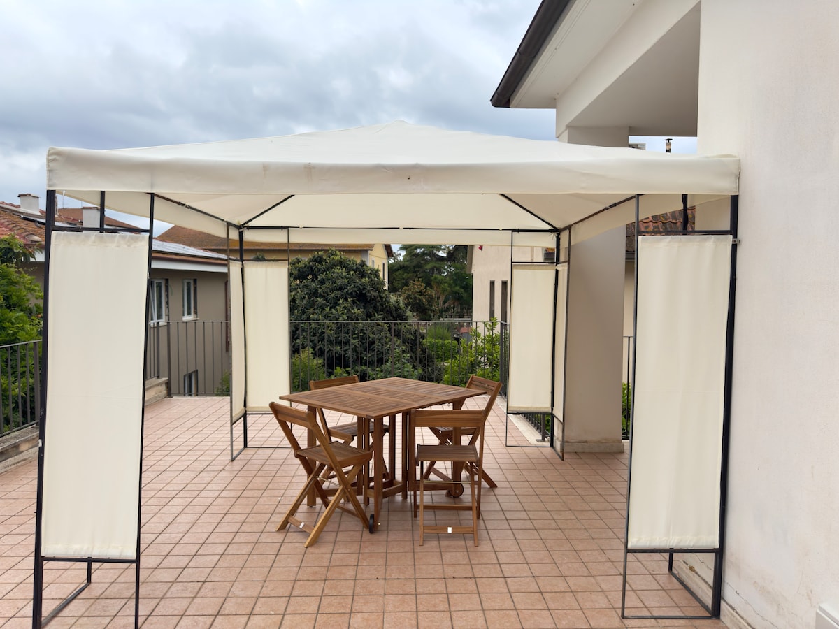 A covered outdoor area features a gazebo with a neutral fabric canopy. Below, a wooden table is surrounded by four folding chairs, providing a space for dining or relaxation. The patio is tiled and offers views of the surrounding greenery.