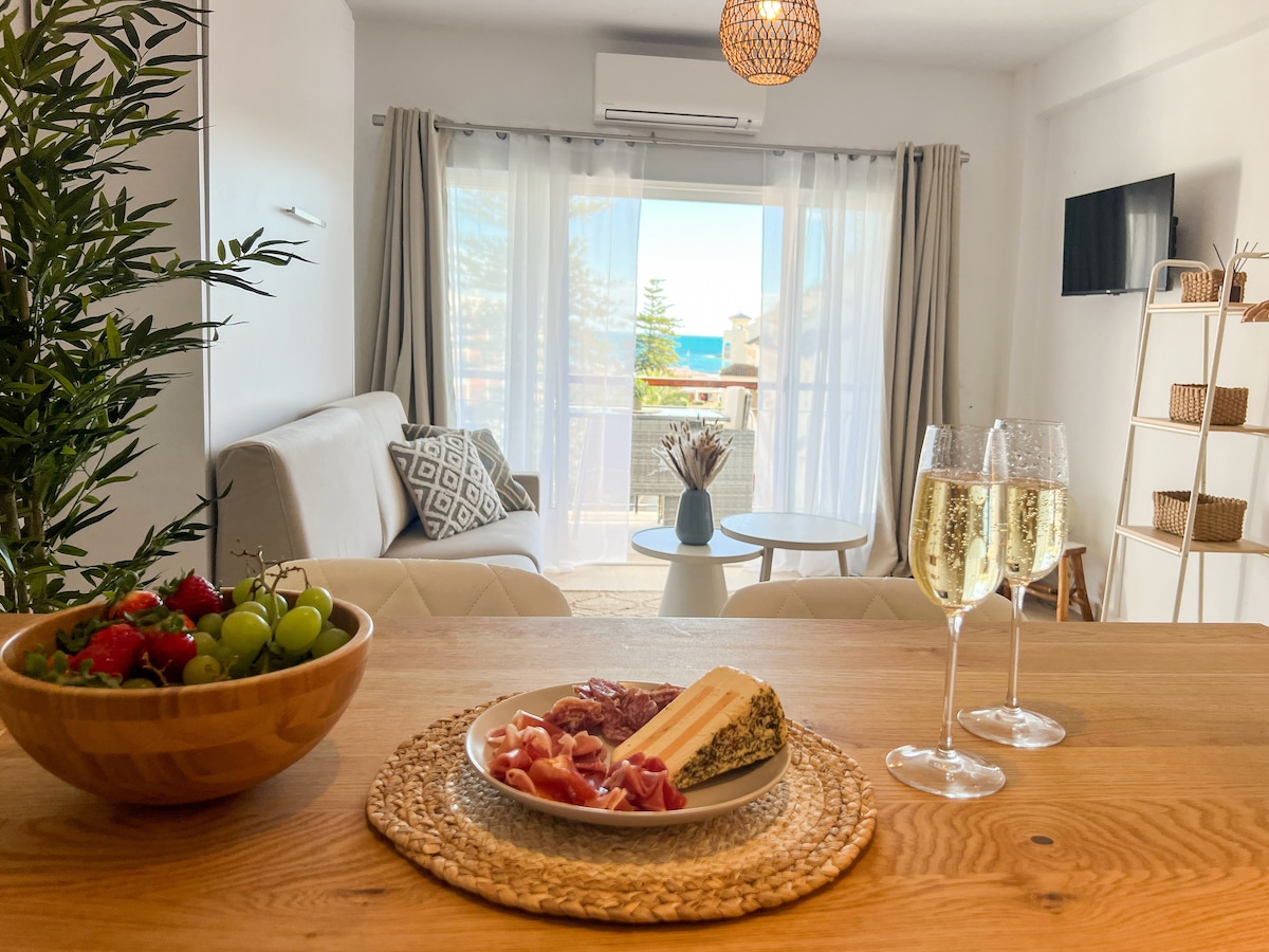 A dining table is set with a plate of assorted cheeses and cured meats, accompanied by fresh grapes and cherry tomatoes. In the background, a spacious living area features a sofa, coffee table, and large windows overlooking the sea, creating a bright and inviting ambiance.