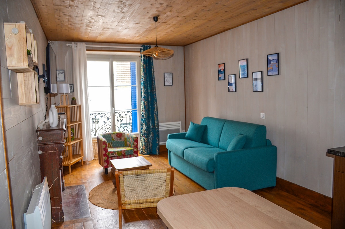 The living area features a comfortable turquoise sofa, a vibrant armchair, and a wooden dining table. Light pours in through large windows, adorned with colorful curtains, while framed pictures decorate the walls. A natural fiber rug enhances the warm, wooden flooring.