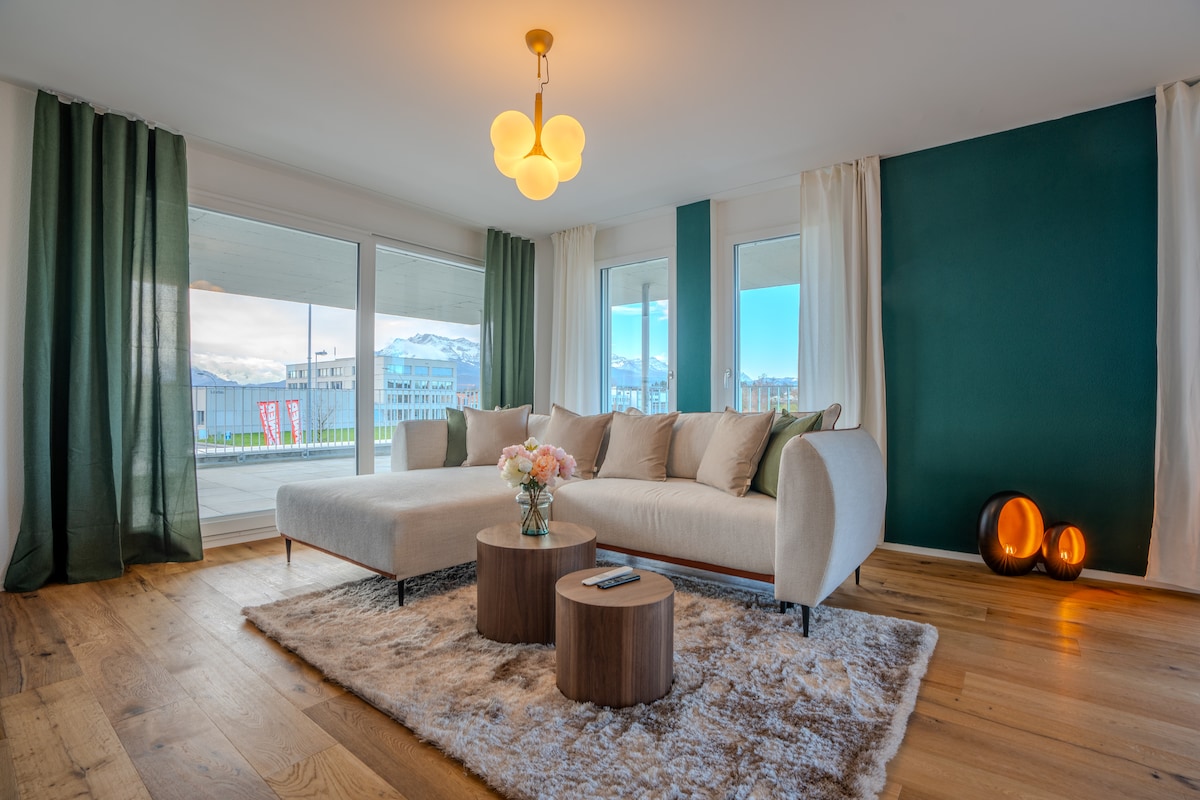 A spacious living area features large windows with mountain views, allowing natural light to fill the room. A light-colored sofa is paired with a set of wooden side tables, placed on a textured rug. Floor-to-ceiling curtains frame the windows, enhancing the overall ambiance.