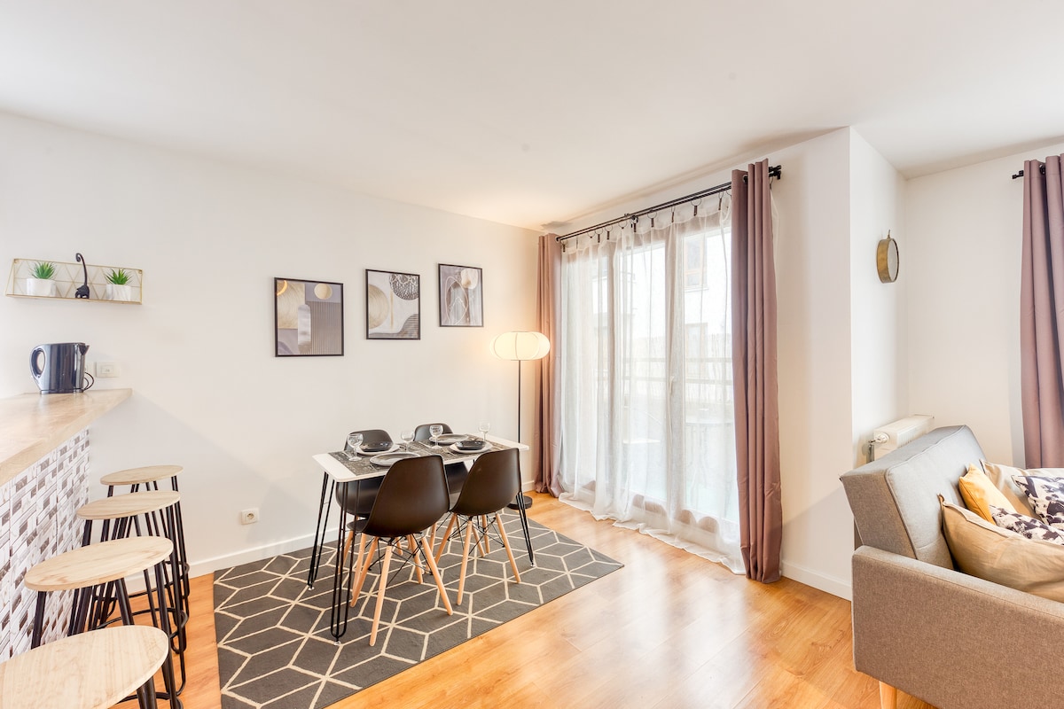 A bright and open living area features a dining table with four chairs and stylish wall art. A comfortable couch is positioned nearby, and natural light flows through large patio doors leading to a balcony, enhancing the inviting atmosphere of the space.