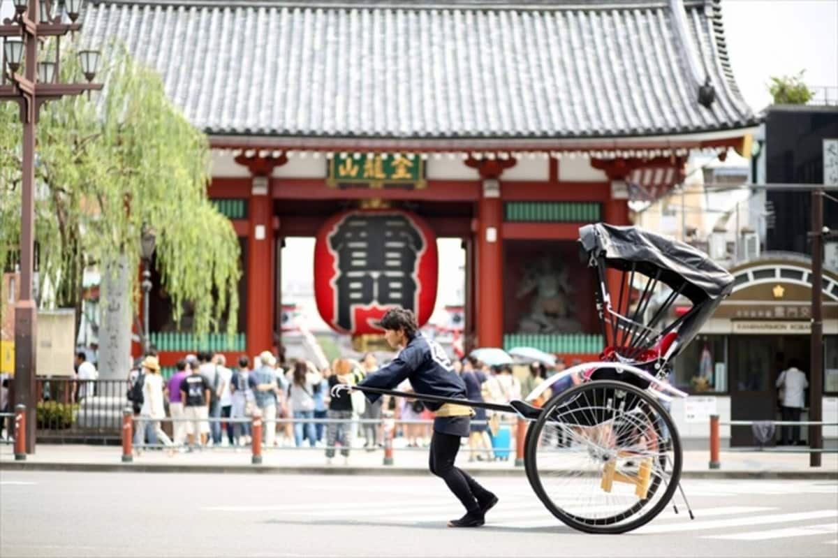 A traditional rickshaw is being pulled along a busy street near a large temple gate adorned with a prominent red lantern. A crowd of visitors can be seen in the background, with lush greenery framing the scene under a clear sky.