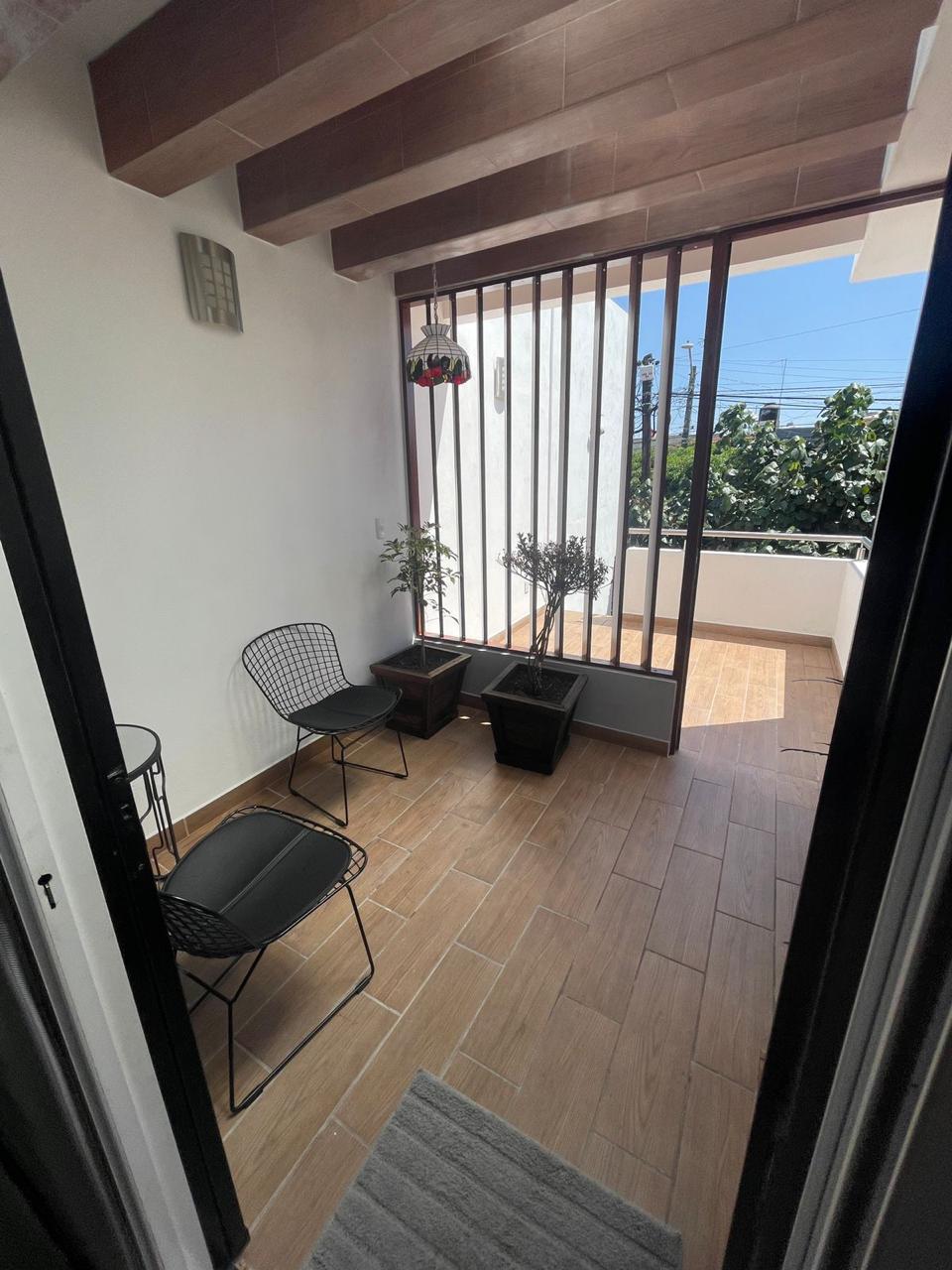 A small balcony area features two black wire chairs positioned nearby several planters. Natural light fills the space through large windows, and a decorative hanging plant adds a touch of greenery. The flooring consists of light-colored tiles, enhancing the open feel.