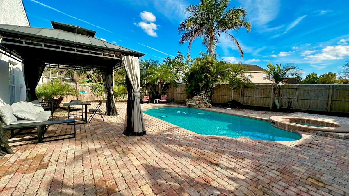 A private outdoor area features a pool surrounded by tropical plants. A gazebo with flowing curtains offers a shaded seating area, complemented by lounge chairs on a brick patio. The clear blue sky is visible above, enhancing the serene ambiance.