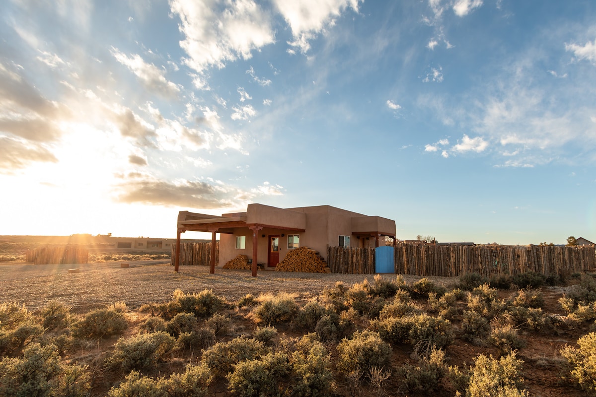 The exterior of a southwestern-style home is presented, featuring a stucco façade with a red door. Sunlight casts a warm glow, illuminating the surrounding sagebrush landscape and wooden fencing. Stacked firewood is visible near the entrance, adding a rustic touch to the serene setting.