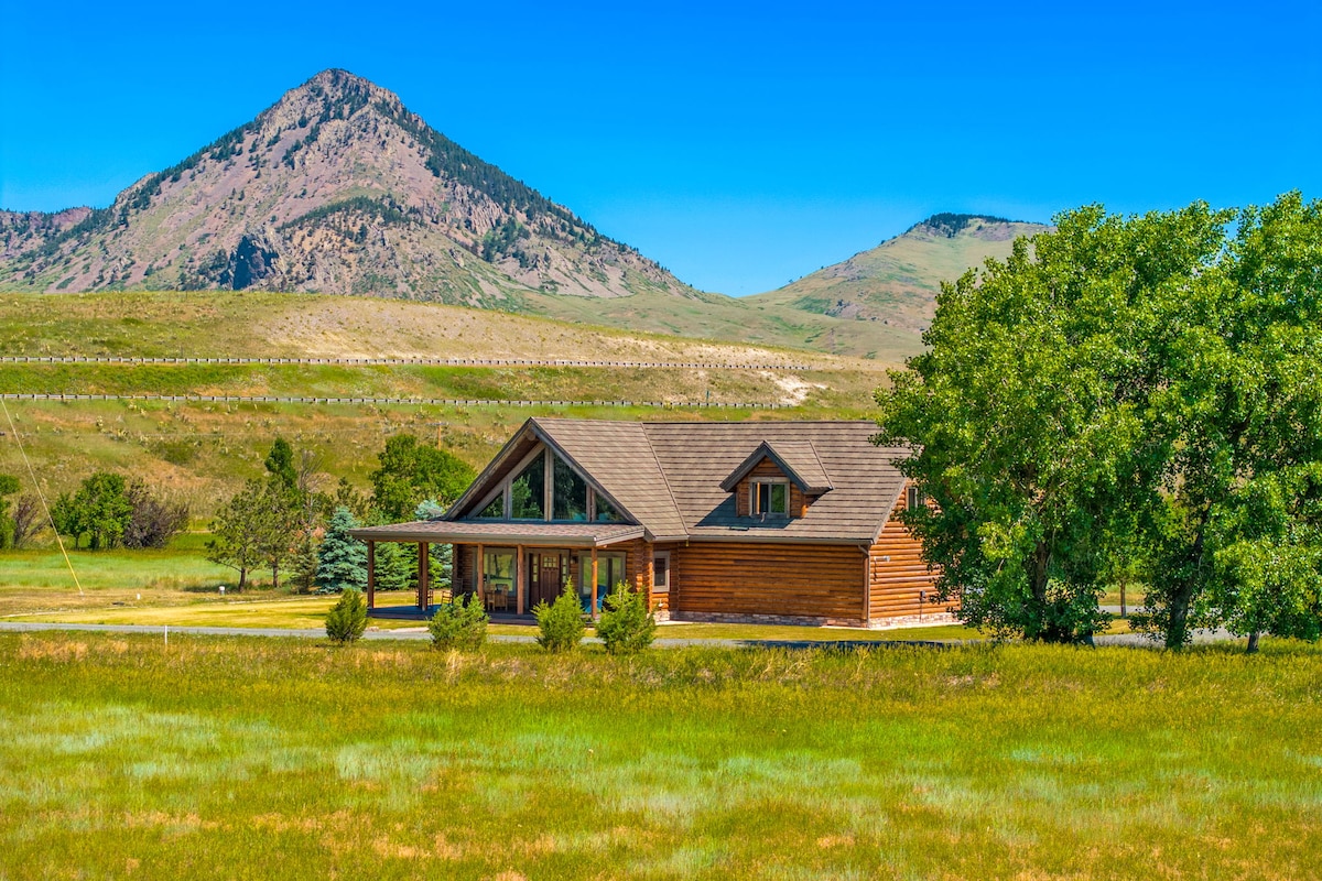 The lodge is set against a backdrop of rolling hills and mountains, featuring a mix of lush green grass and trees. Its wooden exterior is complemented by large windows that bring in natural light and showcase the surrounding landscape.