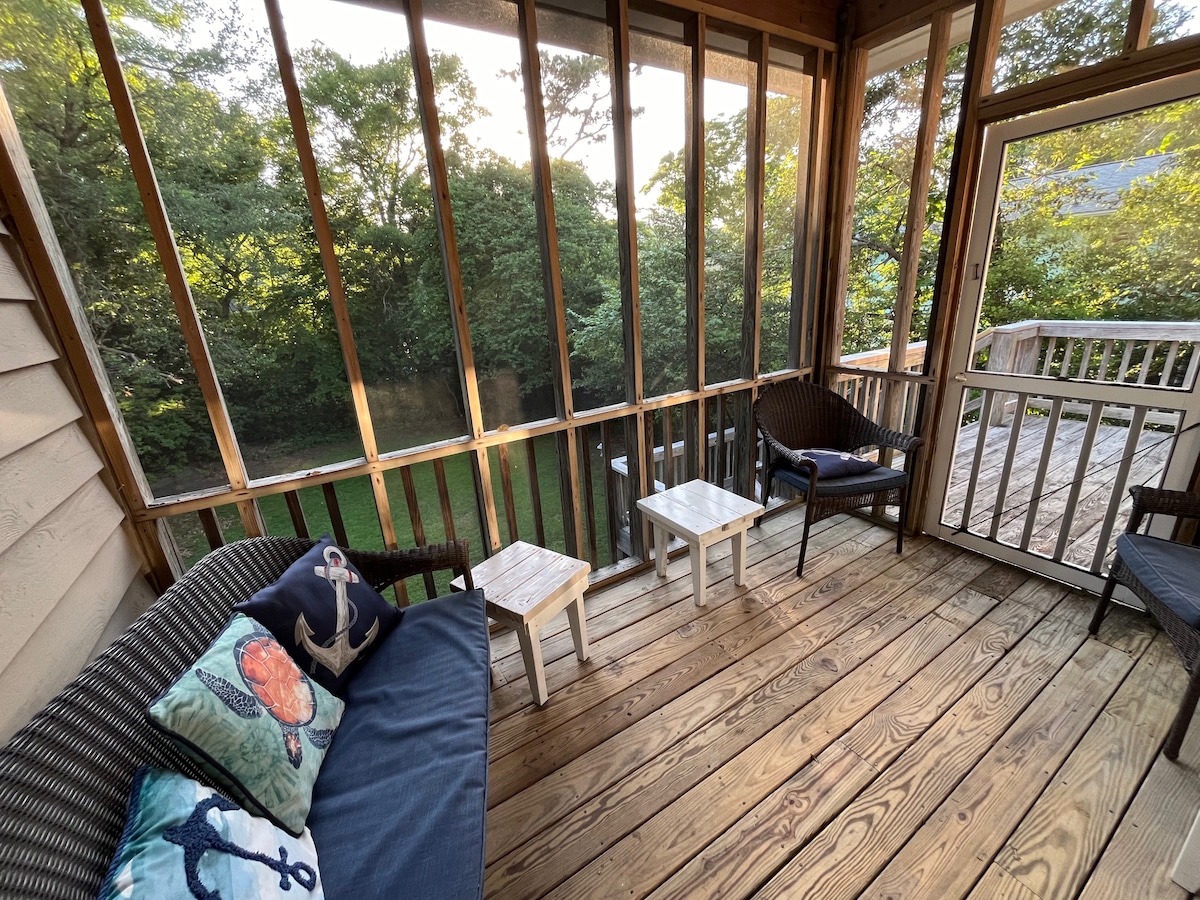 A screened porch features wooden flooring and open framing, providing views of the surrounding greenery. Comfortable seating is arranged, including a dark wicker couch and two small tables, allowing for a relaxing outdoor experience in a private setting.