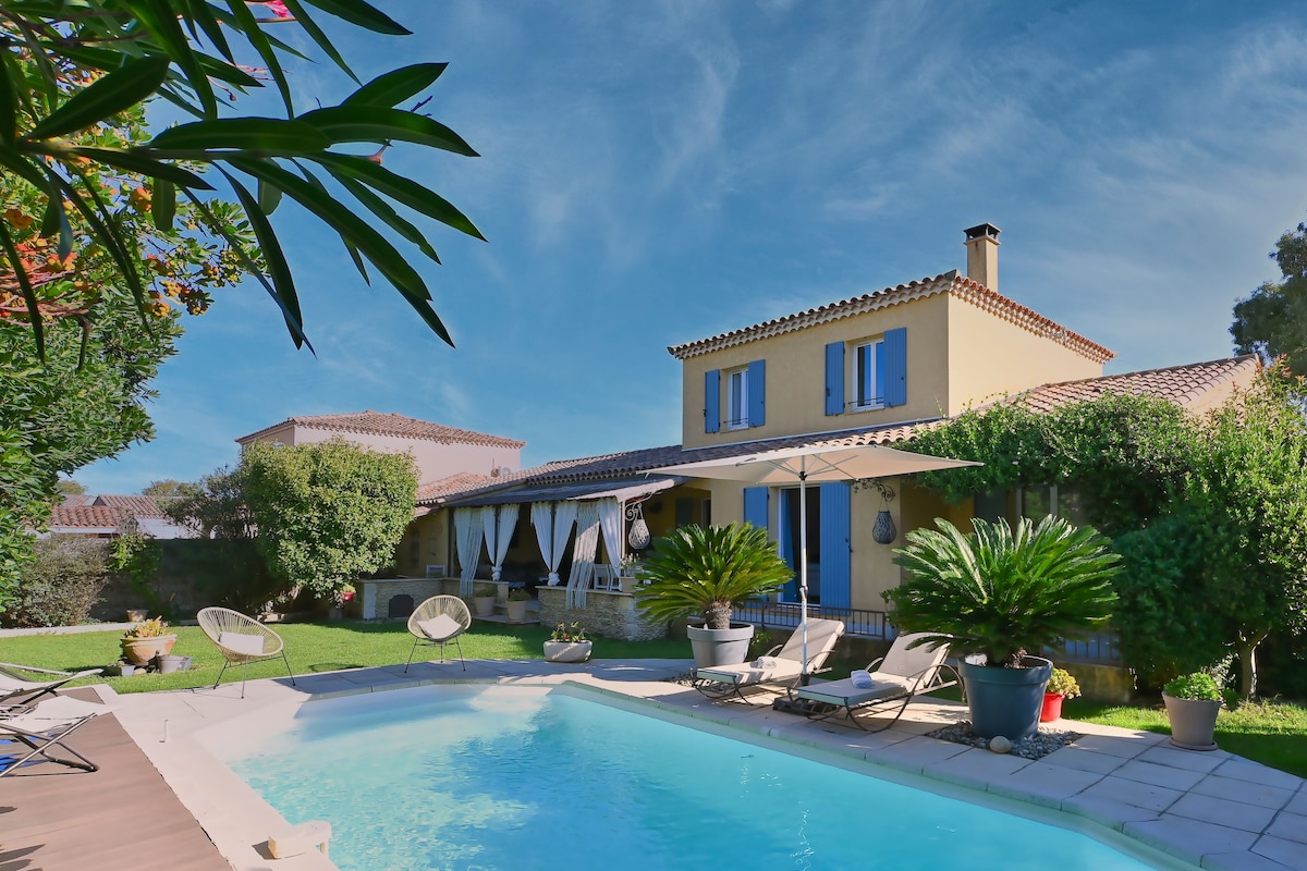 The villa features a sunny exterior with yellow walls and blue shutters, complemented by lush greenery. A private pool with sun loungers sits in the foreground, while a shaded patio area with curtains invites relaxation. The two-story structure offers a cozy and welcoming atmosphere.