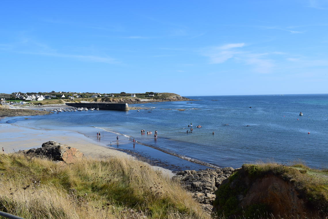 A serene beach scene captures people enjoying the water along a sandy shore. Gentle waves lap against the beach, while rocky formations and lush grass line the coastline. The clear blue sky enhances the peaceful atmosphere, inviting leisurely activities by the sea.
