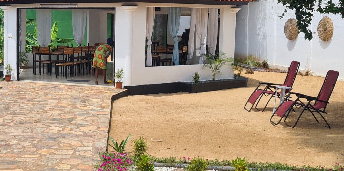An outdoor seating area is presented, featuring two reclining chairs positioned on a stretch of yellow sand. A spacious terrace with large windows overlooks the garden. Potted plants and small shrubs are arranged along the pathway, enhancing the inviting ambiance of the villa.