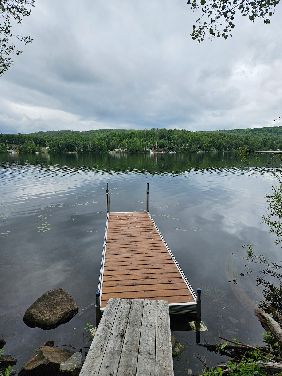 A wooden dock extends into the calm waters of Lac Long, reflecting the surrounding greenery. The shoreline is lined with trees and distant houses can be seen across the lake, creating a serene and natural setting.