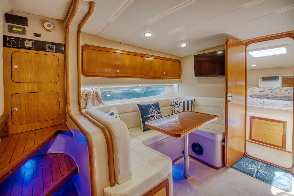 An interior space of a 12-meter boat is shown, featuring light-colored seating around a central table. Nautical-themed cushions decorate the area, while warm wooden cabinetry provides storage. Soft lighting enhances the cozy atmosphere, and a small staircase leads to additional sleeping quarters.