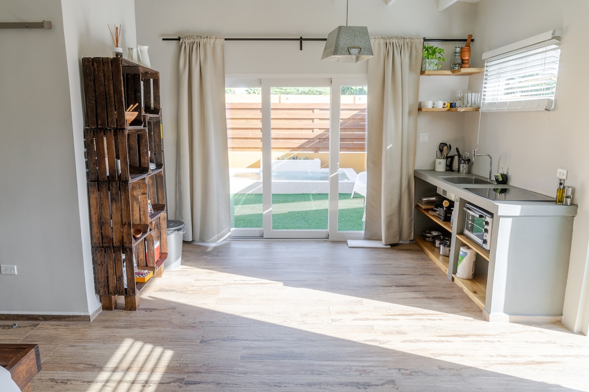 An open living space features wide sliding doors leading to a private outdoor area. Light-colored walls and wooden elements create a calming atmosphere. A compact kitchen with wooden shelving offers essential amenities, while a wooden crate storage unit adds character to the room.