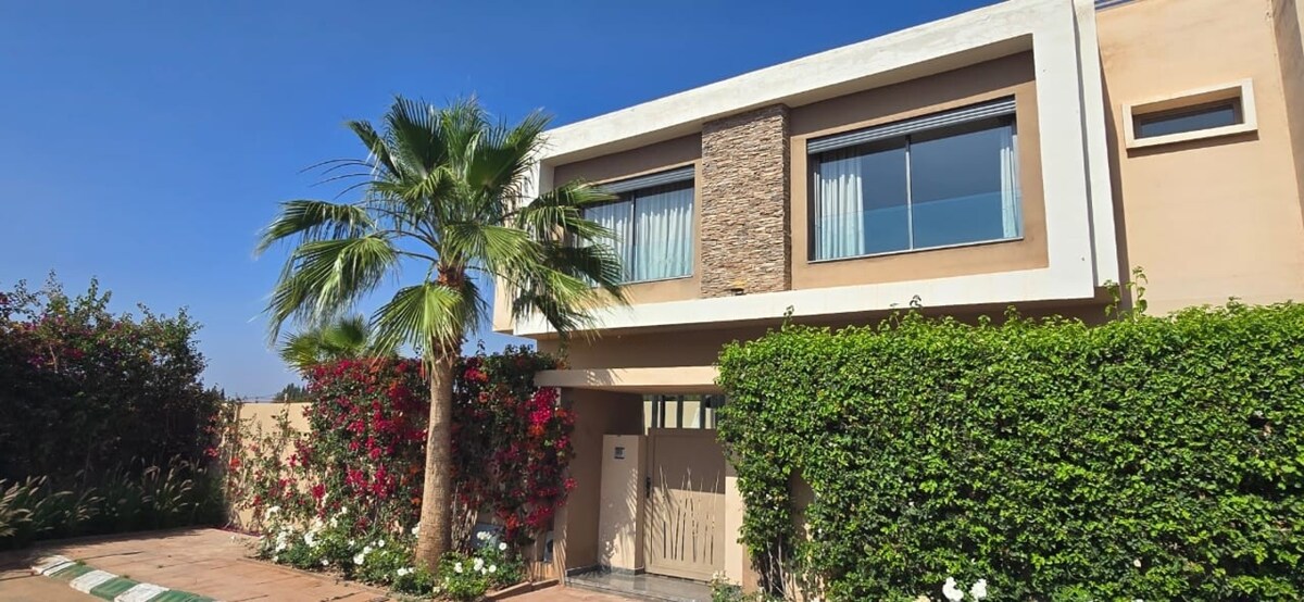 The exterior of the villa is highlighted by lush greenery, with blooming flowers framing the entrance. A tall palm tree provides shade against the clear blue sky, while the modern facade features stone accents and large windows welcoming natural light.