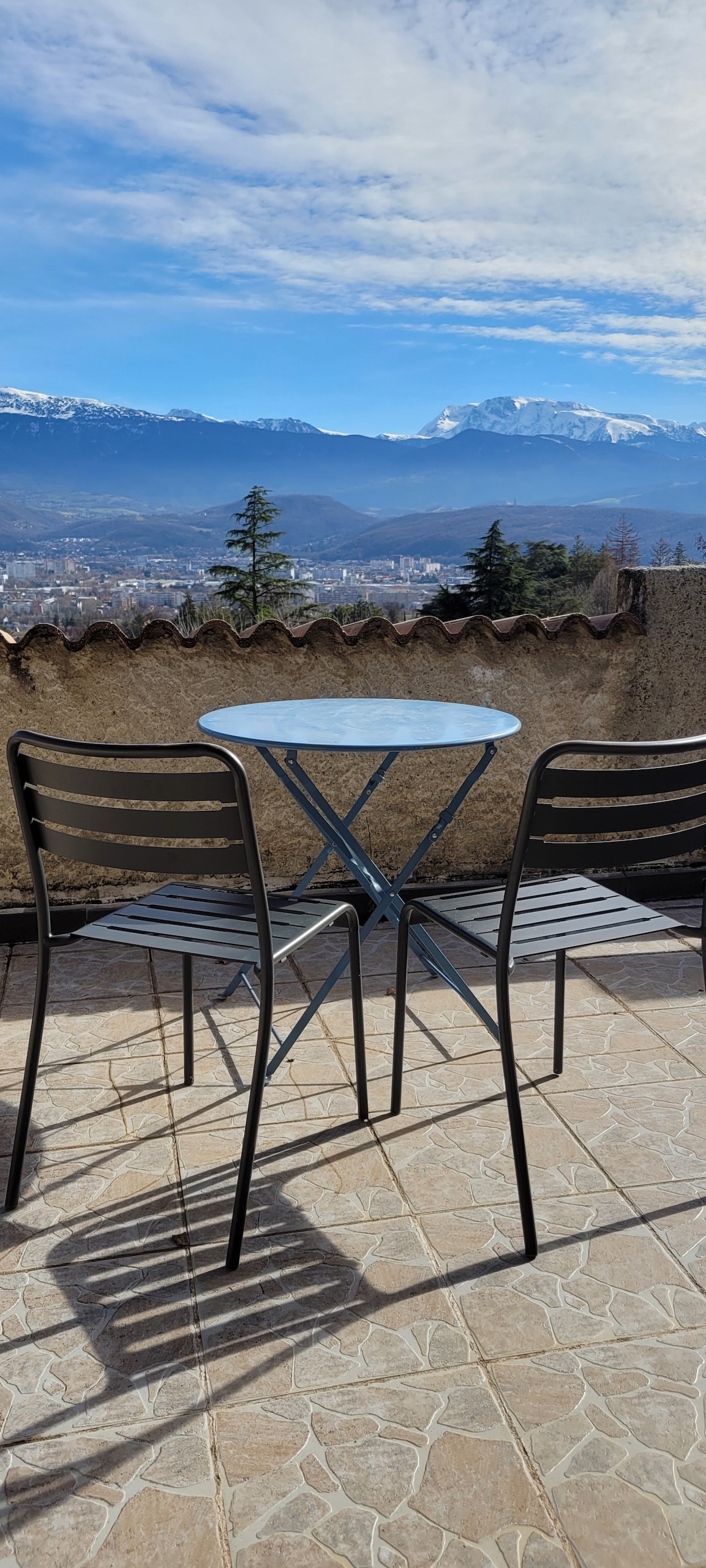 A small terrace is shown with a round metal table and two black chairs, offering unobstructed views of the surrounding mountains and valley. The ground is tiled, and the sky is partly cloudy, framing the scenic backdrop.