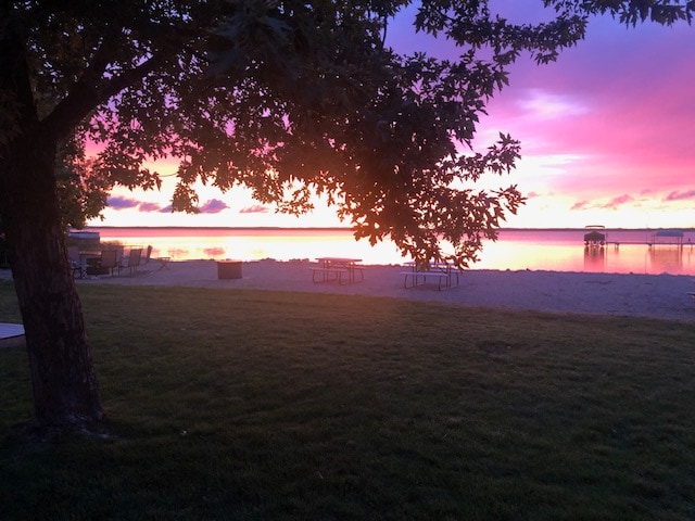 A serene view of the lake is framed by a tree, showcasing vibrant sunset colors reflecting on the water. Picnic tables are visible on the sandy beach, along with a fire pit, creating a peaceful outdoor gathering space.