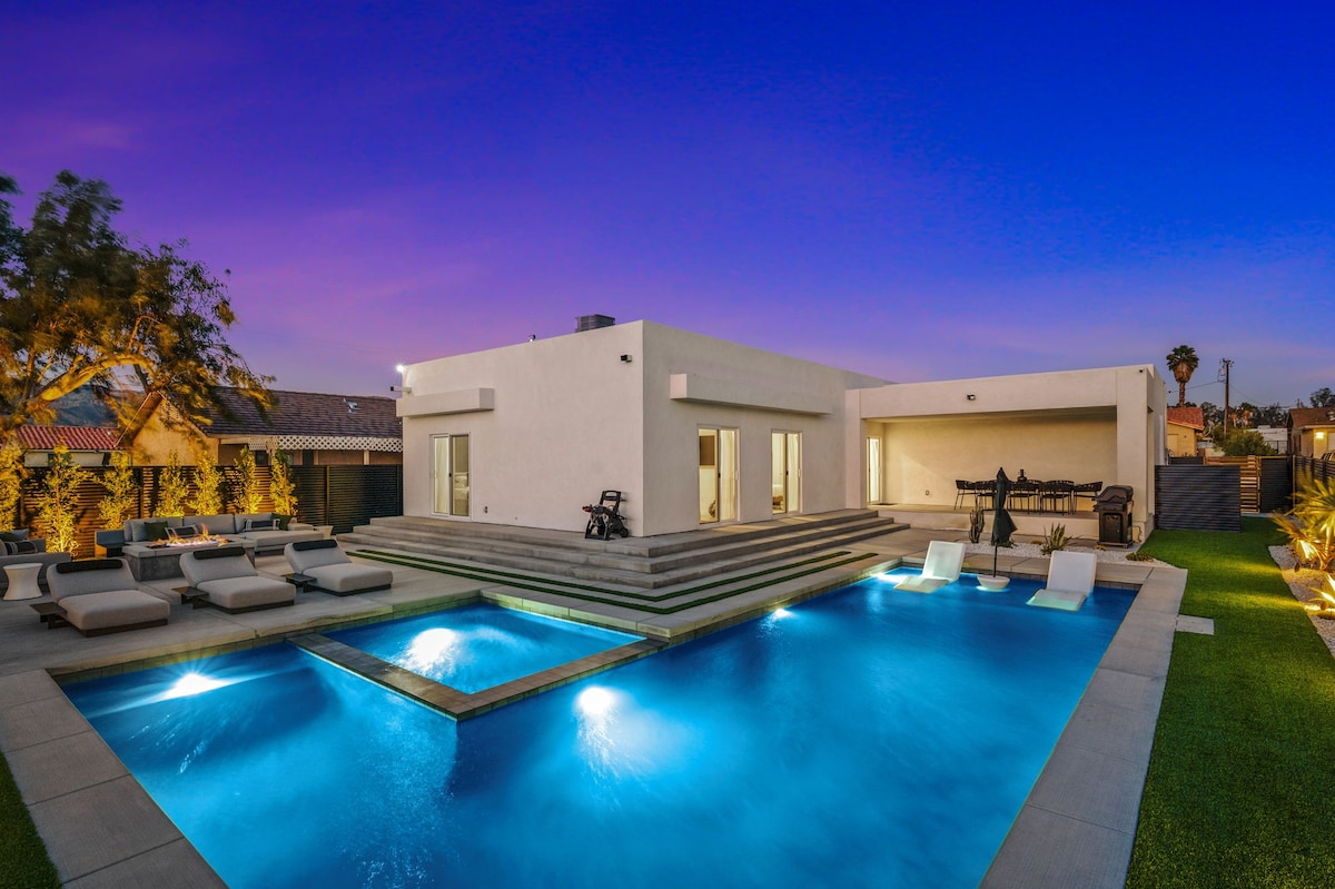 The home's modern architecture is highlighted against a vibrant sunset sky. The outdoor area features a spacious saltwater pool with stepped edges, complemented by lounge chairs and a seating area. Large glass doors provide access to the interior, enhancing the seamless connection to the outdoor space.