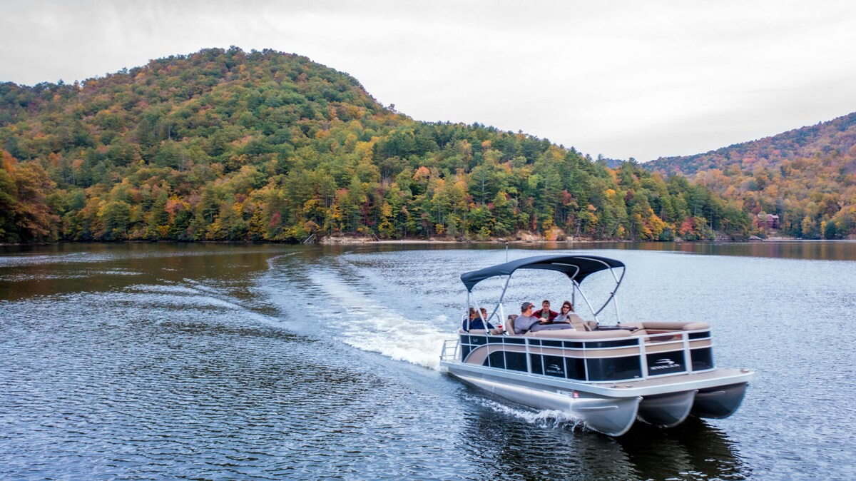 A pontoon boat is shown gliding across a calm lake, surrounded by trees displaying various autumn colors on the distant hills. The boat carries several passengers, enjoying their time on the water while gentle waves trail behind.