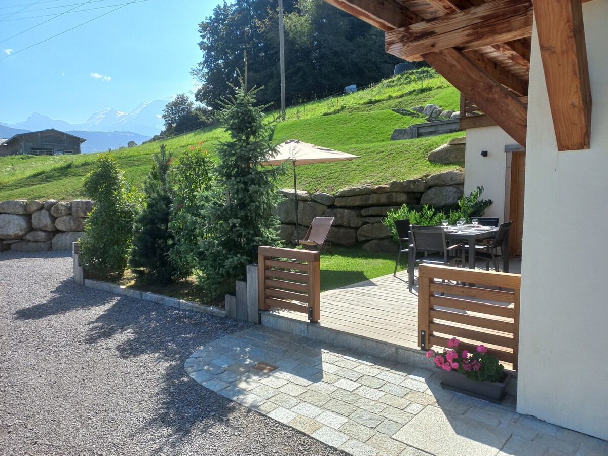 An outdoor area features a wooden deck with seating arrangements, surrounded by greenery and ornamental plants. A large umbrella provides shade, and the distant mountains are visible under a clear blue sky, adding a serene backdrop to the setting.